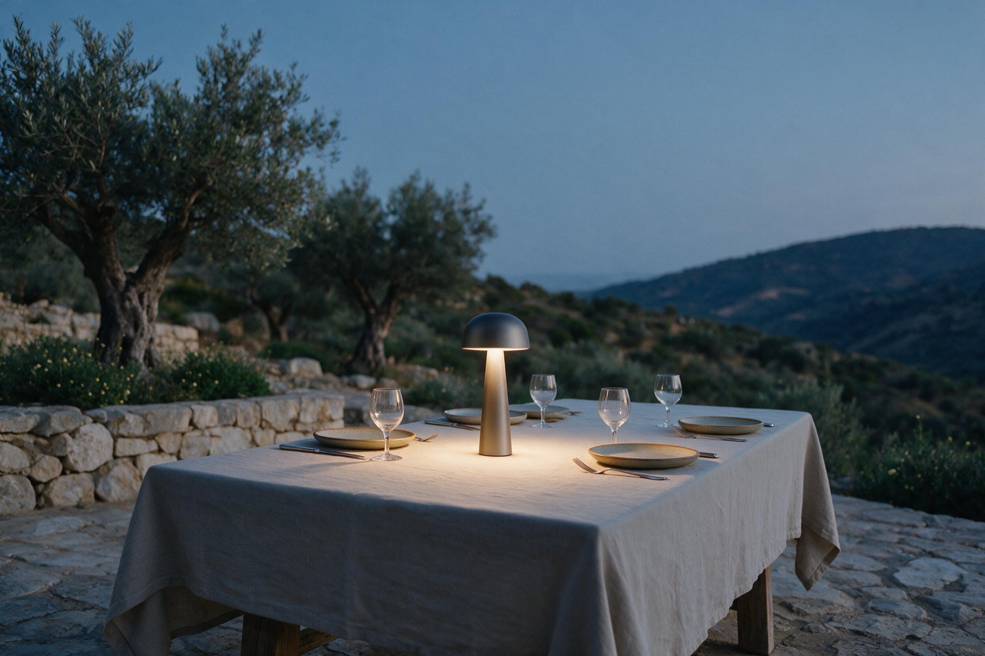 A dinner table set with linen and ceramics in an olive grove at dusk, lit only by the warm glow of the Sera One portable lamp at its center